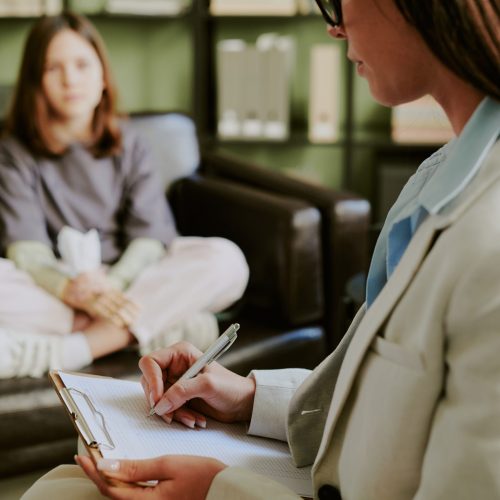 Therapist wearing suit taking notes while counseling young woman sitting on couch in office providing professional support and advice for client's mental health